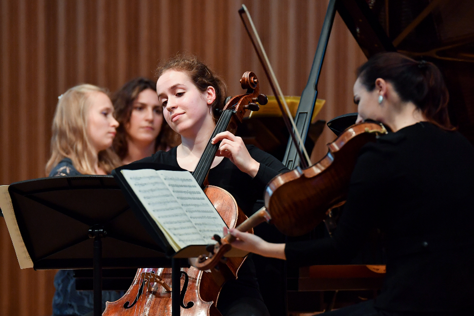A female student, playing on a cello, smiling and reading the music sheet, sitting with other students performing.
